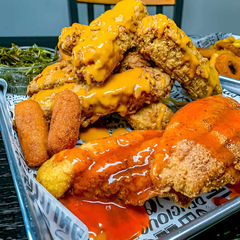 A tray of southern fried chicken tenders with cheese sauce, hot sauce, hush puppies, and collard greens.