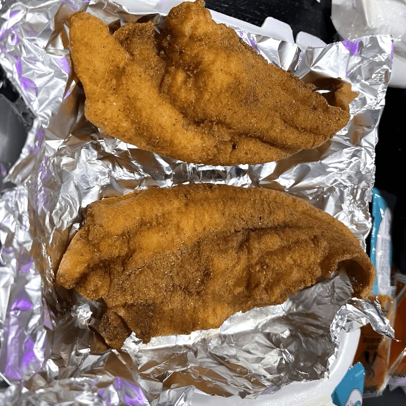 Two crispy, golden-brown fried fish fillets, seasoned with spices, served in aluminum foil.